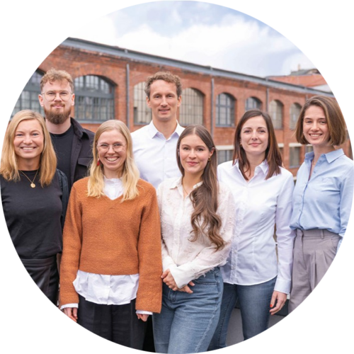 Sieben Personen stehen im Freien vor einem Backsteingebäude, posieren für ein Gruppenfoto und lächeln in die Kamera.