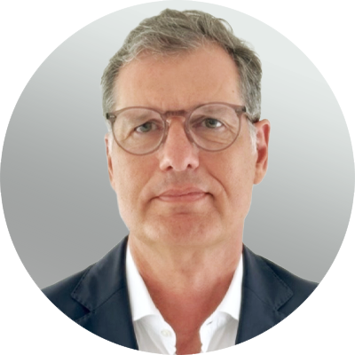 Ein Mann mittleren Alters mit grauem Haar, Brille und neutralem Gesichtsausdruck, der eine dunkle Anzugsjacke und ein weißes Hemd trägt, vor einem hellgrauen Hintergrund.