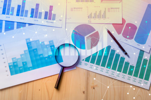 Eine Vielzahl von Finanzdiagrammen und -grafiken sind mit einer Lupe, einem Stift und einer digitalen Datenüberlagerung über einen Tisch verteilt und suggerieren eine detaillierte Analyse.
