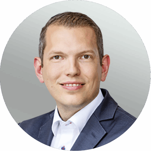 Ein Mann in dunklem Blazer und weißem Hemd lächelt vor hellgrauem Hintergrund in die Kamera.