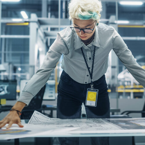 Eine Person mit kurzen, hellen Haaren und Brille, die ein gestreiftes Hemd und einen Ausweis trägt, beugt sich über einen Tisch und betrachtet einen großen Bauplan in einem modernen, industriell anmutenden Büro.