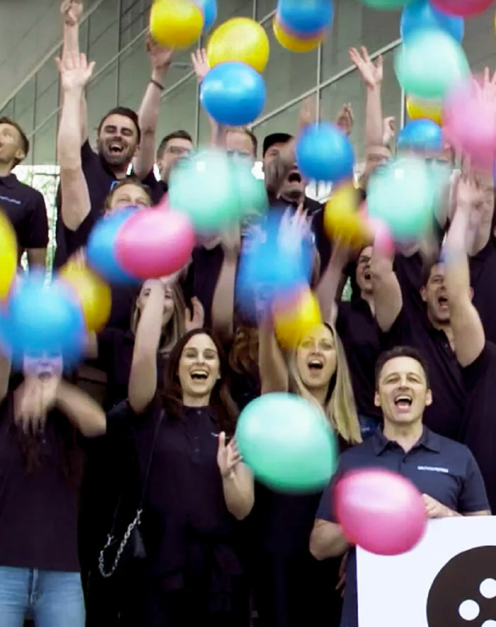 Eine Gruppe von Menschen in dunklen Hemden steht lächelnd und jubelnd vor einem Gebäude, während bunte Luftballons in die Luft geworfen werden.