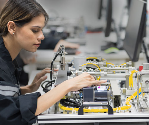 Eine Frau arbeitet mit elektronischen Komponenten und Maschinen an einer Werkbank in einem Labor oder Industrieumfeld.