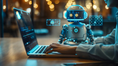 Ein kleiner, humanoider Roboter mit blauen Digitalanzeigen steht neben einer Person, die auf einem Laptop tippt, vor einem verschwommenen Lichthintergrund in einer Innenumgebung.