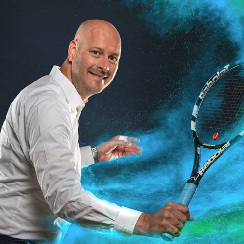 Eine Person in einem weißen Hemd hält einen Tennisschläger vor einem Hintergrund aus blau und grün gefärbtem Rauch oder Pulver.