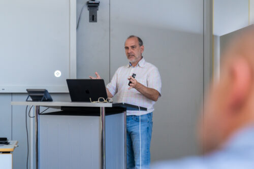 Ein Mann in Freizeithemd und Jeans steht an einem Podium und hält eine Präsentation. Im Hintergrund sind ein Laptop und eine Projektionsleinwand zu sehen.