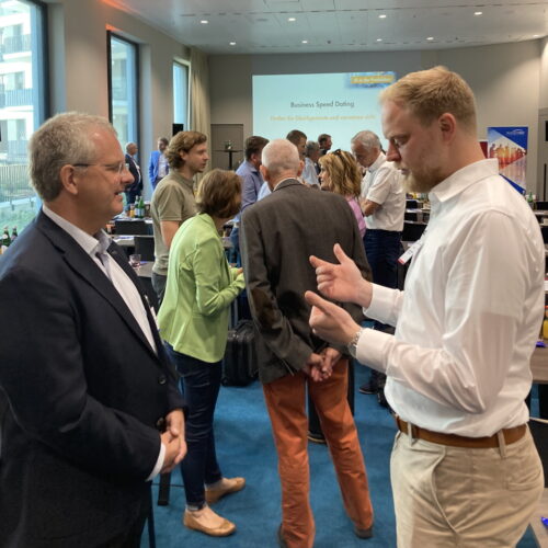 Bei einer Networking-Veranstaltung unterhalten sich Menschen. Im Vordergrund diskutieren zwei Männer, im Hintergrund sind weitere Teilnehmer neben einem Präsentationsbildschirm zu sehen.