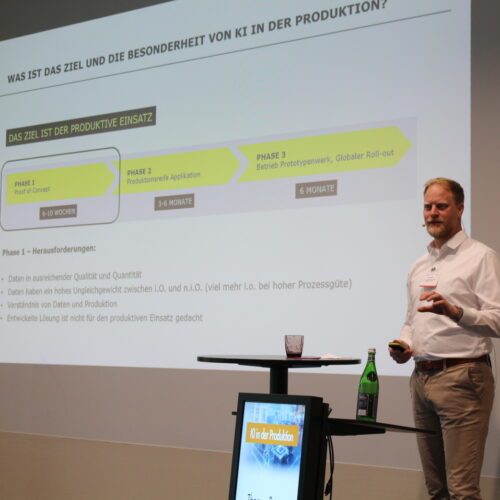 Ein Mann präsentiert vor einem Bildschirm einen schrittweisen Projektplan auf Deutsch. Die Folie diskutiert den Einsatz künstlicher Intelligenz in der Produktion und enthält einen Zeitplan und damit verbundene Herausforderungen.