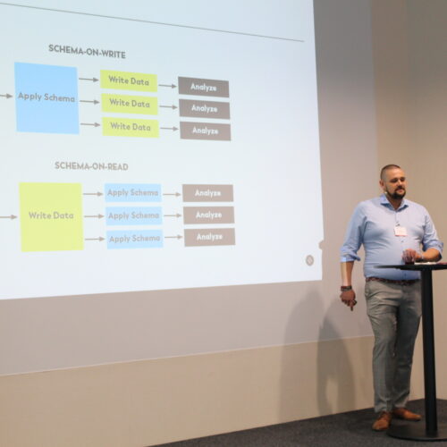 Eine Person steht neben einer Projektionsfläche und erklärt anhand von zwei Flussdiagrammen die Unterschiede zwischen Schema-on-Write und Schema-on-Read.