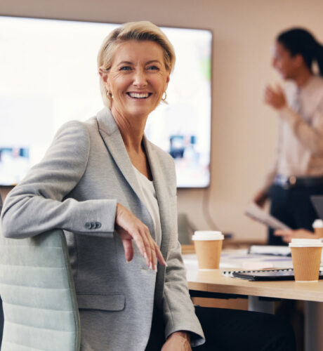 Eine Frau im grauen Blazer sitzt lächelnd an einem Schreibtisch mit Kaffeetassen und Papierkram, während im Hintergrund eine weitere Person neben einem Bildschirm steht.