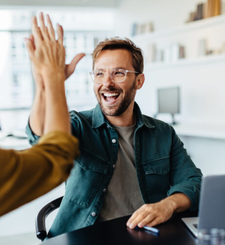 Ein Mann mit Brille und grüner Jacke lächelt und gibt einer anderen Person im Haus ein High-Five. Auf dem Tisch vor ihm ist ein Laptop zu sehen.