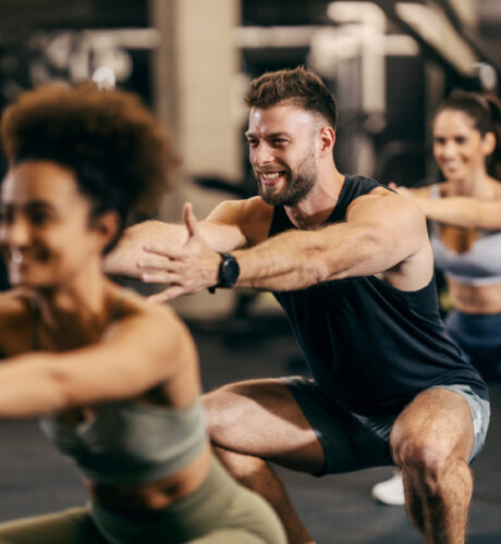 Eine Gruppe von Menschen trainiert in einem Fitnessstudio und konzentriert sich auf Kniebeugen. Der Mann vorn lächelt und alle tragen Sportkleidung.