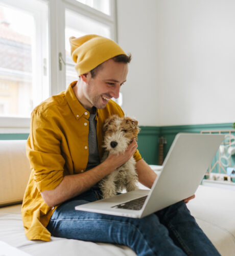 Ein Mann in einem gelben Hemd und einer gelben Beanie-Mütze sitzt auf einem weißen Sofa, hält einen kleinen Hund und lächelt auf einen Laptop-Bildschirm.