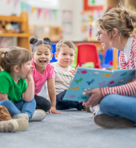 Eine Frau liest drei kleinen Kindern, die in einem Klassenzimmer auf dem Boden sitzen, aus einem bunten Buch vor. Alle Kinder sind aufmerksam und lächeln.