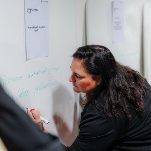 Innovators Club. Eine Person schreibt während eines Workshops mit einem Marker auf ein Whiteboard. Über der Schreibfläche hängt ein Zettel mit der Aufschrift „INNOVATORS CLUB“ und einer Frage in deutscher Sprache.