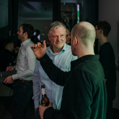 Innovators Club. Mehrere Personen unterhalten sich drinnen. Ein Mann mit grauem Haar unterhält sich mit einem Glatzkopf, der eine Bierflasche hält. Andere Personen unterhalten sich im Hintergrund.