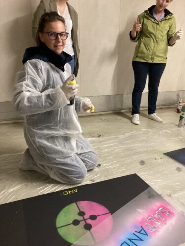 Eine Person in Schutzanzug und Handschuhen kniet und hält eine Sprühdose in der Hand. Sie posiert neben einem farbenfrohen Kunstwerk mit wissenschaftlichem Thema auf dem Boden. Zwei weitere Personen stehen im Hintergrund.