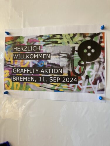 Ein Plakat mit buntem Graffiti-Hintergrund und dem Text „Herzlich Willkommen Graffiti-Aktion Bremen, 11. Sep 2024“, befestigt an einem Whiteboard.