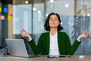 Eine Frau im grünen Blazer sitzt mit einem Laptop an einem Schreibtisch, die Augen sind geschlossen und die Hände in einer Meditationspose.