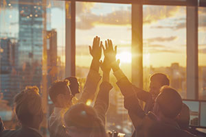 Eine Gruppe von Menschen in einem Büro gibt sich ein High-Five, während das Sonnenlicht durch die großen Fenster strömt.