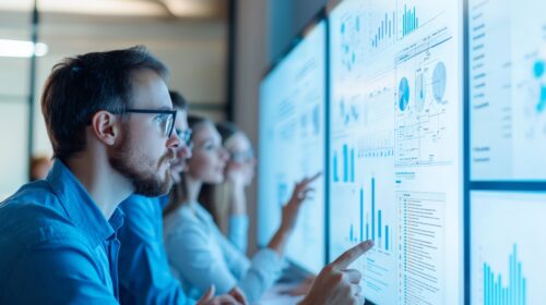Vier Personen analysieren in einer Büroumgebung Diagramme und Datentabellen auf großen digitalen Bildschirmen.