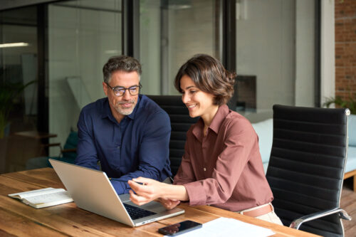 Zwei Personen sitzen an einem Schreibtisch in einem Büro, schauen auf den Bildschirm eines Laptops und besprechen etwas, auf dem Tisch liegen Notizbücher und ein Telefon.