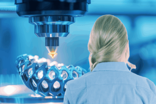 Eine Frau mit blondem Haar beobachtet einen großen 3D-Drucker, der in einer blau beleuchteten High-Tech-Umgebung ein komplexes geometrisches Objekt herstellt.
