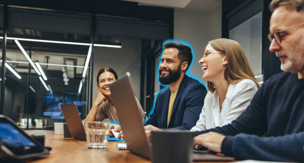 Vier Personen sitzen an einem Konferenztisch mit Laptops, lächeln und diskutieren; ein Mann ist mit einem blauen Umriss hervorgehoben.