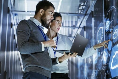 Zwei Fachleute analysieren in einem Rechenzentrum digitale Informationen auf einem großen Bildschirm; einer hält einen Laptop, während der andere auf das Display zeigt.