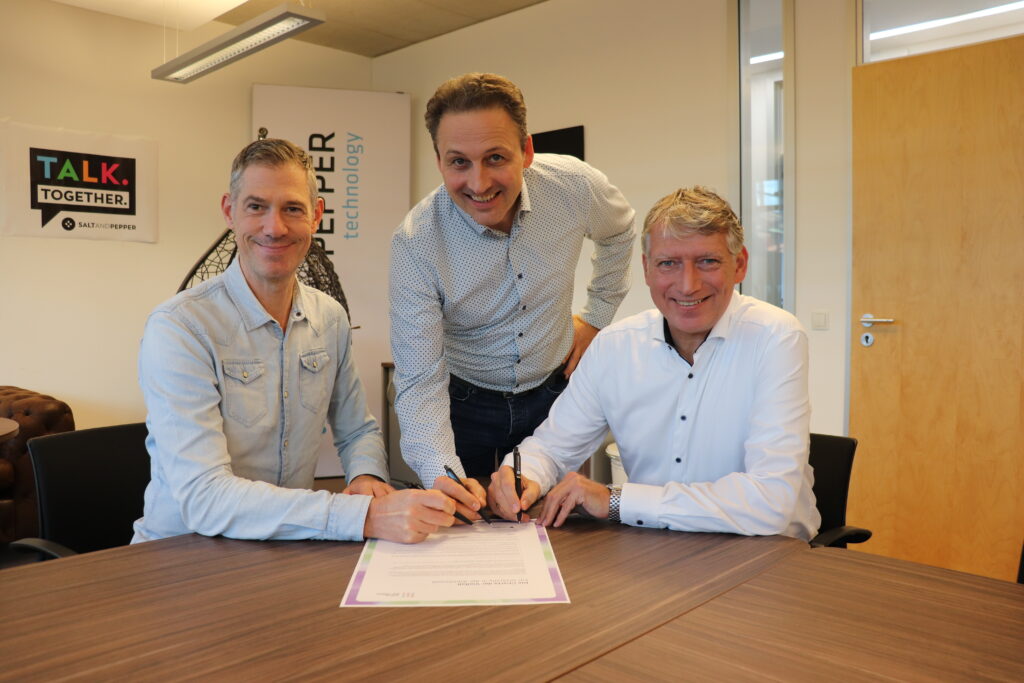 Drei Männer sitzen und stehen an einem Tisch und lächeln, während sie in einem modernen Büro ein Dokument unterschreiben.