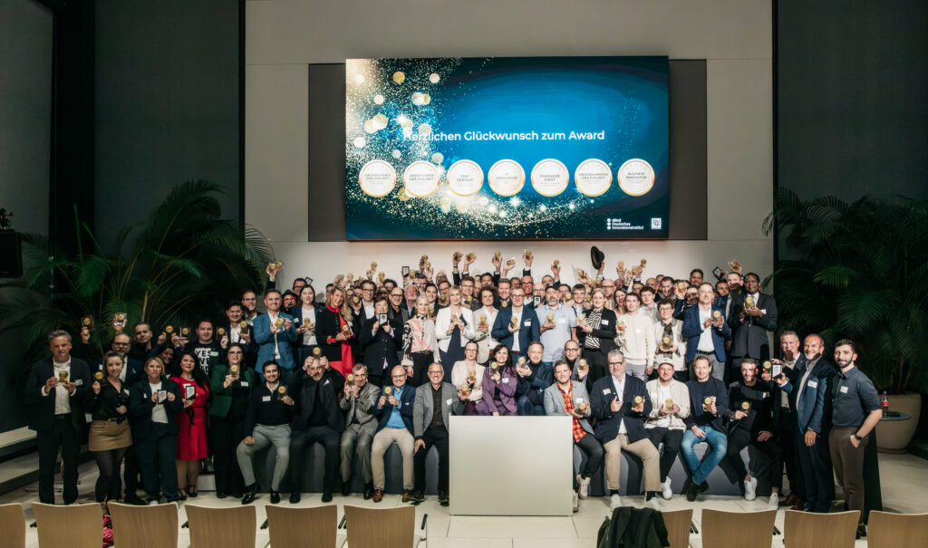 Eine große Gruppe formell gekleideter Menschen posiert für ein Gruppenfoto in einem Innenraum, viele von ihnen mit Trophäen in der Hand, vor einem Bildschirm, auf dem ein Glückwunschtext auf Deutsch angezeigt wird.