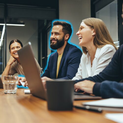 Drei Personen sitzen an einem Konferenztisch mit Laptops, Notebooks und Brillen, lächeln und sind in eine Besprechung in einer modernen Büroumgebung vertieft.