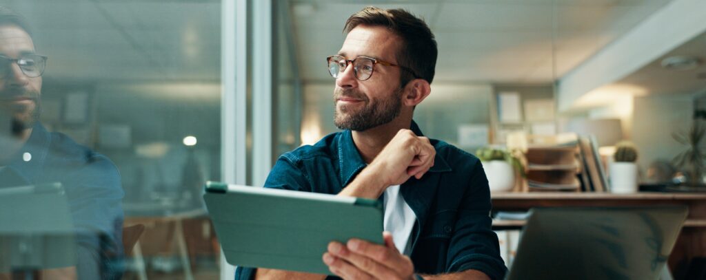 Ein Mann mit Brille sitzt an einem Schreibtisch, hält ein Tablet in der Hand und blickt nachdenklich aus dem Fenster einer modernen Büroumgebung.