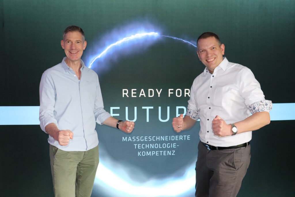 Zwei Männer stehen vor einer Kulisse mit den Aufschriften READY FOR FUTURE" und MASSGESCHNEIDERTE TECHNOLOGIE-KOMPETENZ", beide lächeln und posieren mit geballten Fäusten.